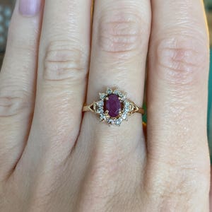 May include: A gold ring with an oval ruby centre stone, encircled by smaller, clear gemstones. The ring's band has a simple, delicate design. The ring is set on a hand.
