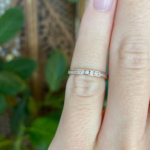 May include: A gold band ring with a row of small, clear gemstones. The ring is worn on a finger, with the skin tone appearing light. The background is blurred, with green foliage and a glimpse of a wooden surface.