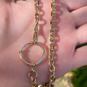 May include: A gold-colored necklace with a large circular pendant and a chain link design. The necklace is made of a shiny, reflective metal and is displayed against a neutral background. The necklace is a jewelry item.