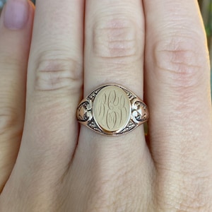 May include: A gold signet ring with an oval face featuring engraved initials. The ring has decorative filigree on the shoulders and a polished finish. The ring is a classic piece of jewelry.