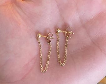 May include: A pair of gold chain earrings. Each earring features a small ball stud connected to a delicate chain that dangles downwards. The earrings are displayed against a neutral background, highlighting their simple elegance.