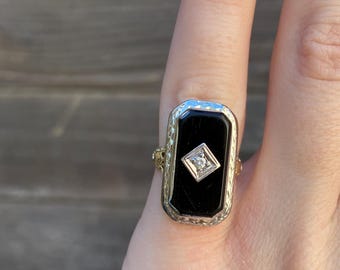 May include: A gold ring with a black onyx stone and a small diamond in the center. The ring has a detailed design around the stone.