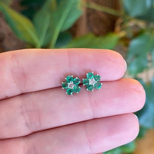 May include: A pair of flower-shaped stud earrings. Each earring features five green gemstones arranged around a central, smaller, clear gemstone. The earrings are set in a silver-colored metal and are displayed against a blurred background.