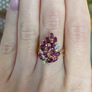 May include: A gold ring with a cluster of oval-cut, deep red gemstones and small, clear accent stones. The ring's design is a stylized floral or leaf shape, set against a gold band. The ring is shown on a person's finger.