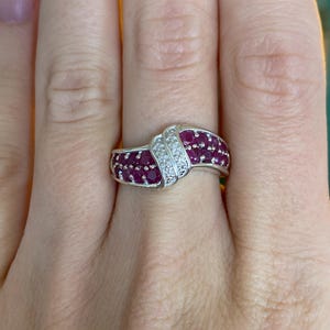 May include: A silver ring featuring a band of small, round, deep red gemstones. The ring has a curved design with a section of smaller, clear gemstones. The ring is on a person's finger.