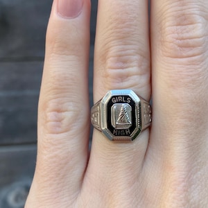 May include: A silver class ring with a black enamel inset. The ring features the words "Girls High" and a stylized pine tree.