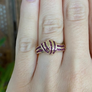 May include: A gold ring featuring a knot design, adorned with rows of small, rectangular, deep red gemstones. The ring is set on a finger, showcasing the intricate details and the contrast between the gold and the gemstones.