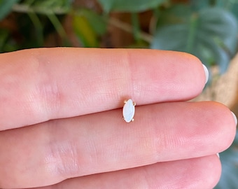 May include: A small marquise-shaped stud earring featuring a white iridescent stone set in a gold-toned metal prong setting. The piece is held between two fingers, showcasing its elongated, pointed oval design and subtle reflective surface.