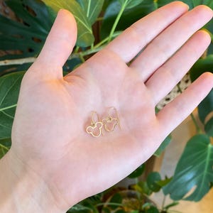 May include: A pair of gold-colored wire earrings in the shape of a mouse head, resting on a person's open palm. The earrings have a simple, minimalist design. The background features green foliage.
