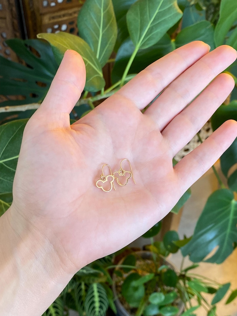May include: A pair of gold-colored wire earrings in the shape of a mouse head, resting on a person's open palm. The earrings have a simple, minimalist design. The background features green foliage.