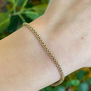 May include: A gold-colored chain bracelet with a delicate, woven design. The bracelet is worn on a wrist, with green foliage in the background. The bracelet's links are closely connected, creating a smooth, continuous appearance.