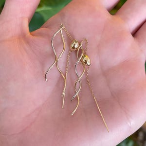 May include: A pair of gold earrings with a delicate design. One earring features a wavy, elongated shape, while the other has a small gold ball and a chain. The earrings are displayed on a hand.