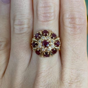 May include: A gold ring featuring a floral design. The ring is adorned with numerous deep red gemstones and small, clear accent stones. The gemstones are arranged in a circular pattern, creating a flower-like appearance. The ring is set on a gold band.