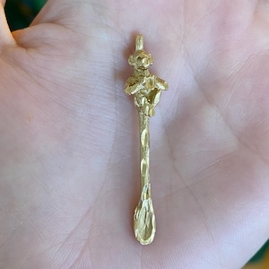 May include: A gold-colored spoon pendant with a monkey figure at the top. The spoon has a textured handle and a small, rounded bowl. The monkey is detailed with facial features and is holding a small object. The pendant is held in a person's hand.