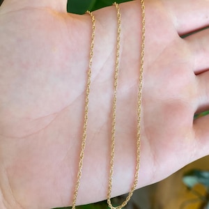 May include: A thin gold rope chain necklace featuring a twisted metal link design. The delicate chain is composed of small, interlocking segments that create a continuous, textured, and reflective surface throughout the entire length of the jewelry piece.