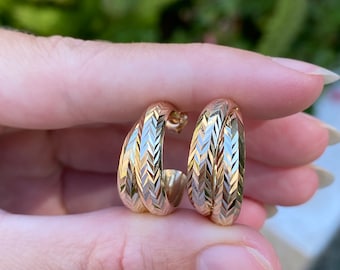 May include: A pair of gold-colored hoop earrings with a textured chevron pattern. The earrings are held up close, showcasing their design and the light reflecting off the surface. The earrings are a classic jewelry item.