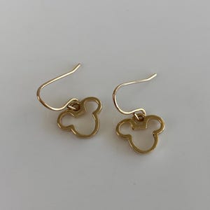 May include: A pair of gold-colored earrings featuring a Mickey Mouse head silhouette. The earrings have a simple hook design and are set against a white background. The earrings are a classic and recognizable design.
