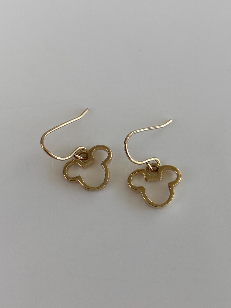 May include: A pair of gold-colored earrings featuring a Mickey Mouse head silhouette. The earrings have a simple hook design and are set against a white background. The earrings are a classic and recognizable design.