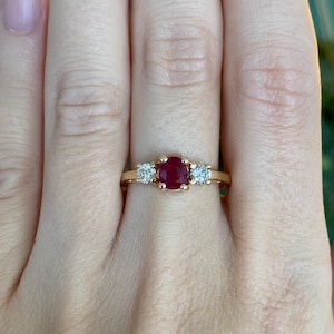 May include: A gold ring featuring a central oval-cut ruby gemstone flanked by two smaller, round, clear gemstones. The ring is set on a thin, gold band, showcasing a classic three-stone design.