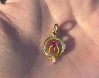 May include: A gold pendant with a red gemstone in the center, surrounded by a circle of gold. A smaller diamond-shaped gemstone hangs from the bottom of the pendant.