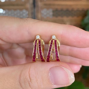 May include: A pair of gold hoop earrings featuring two parallel rows of channel-set square-cut red rubies. Each earring is topped with a single round brilliant-cut white diamond set in a gold bezel at the apex of the arch.