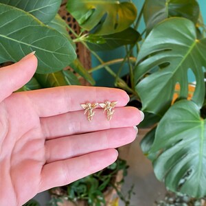 May include: A pair of gold-colored angel earrings. Each earring features a detailed design of an angel with outstretched wings. The earrings are small and delicate, suitable for everyday wear. The earrings are displayed against a backdrop of green foliage.