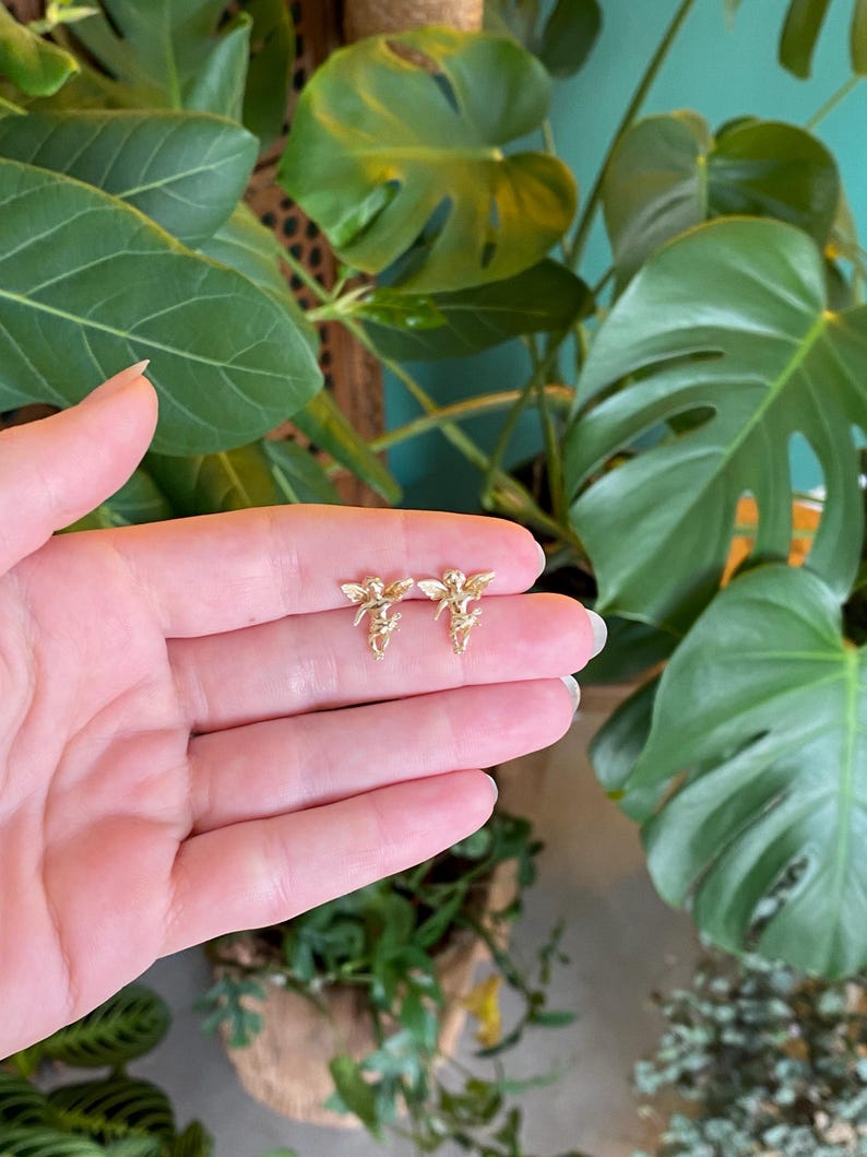 May include: A pair of gold-colored angel earrings. Each earring features a detailed design of an angel with outstretched wings. The earrings are small and delicate, suitable for everyday wear. The earrings are displayed against a backdrop of green foliage.