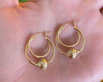 May include: A pair of gold hoop earrings. Each earring features a double hoop design with a textured, spherical bead at the center. The earrings are displayed against a blurred background.