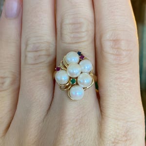 May include: A gold ring featuring a cluster of seven white pearls. The ring is accented with small red, blue, and green gemstones. The ring is on a finger.