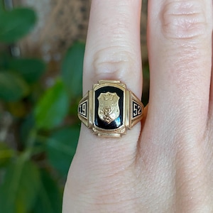 May include: A gold ring with a black central panel displaying a shield emblem. The ring is flanked by the numbers '49' and '52'. The ring is worn on a finger.