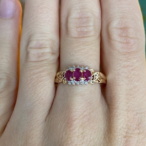 May include: A gold ring featuring three oval-cut, deep red gemstones, possibly rubies, accented with small, clear stones. The ring has an ornate, filigree design on the band and around the central stones. The ring is displayed on a person's finger.