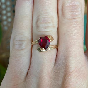 May include: A gold ring with a heart-shaped, deep red gemstone. The ring's band has a delicate, swirling design with small gold accents near the gemstone. The ring is shown on a person's finger.