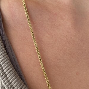 Puede incluir: Un collar de cadena de cuerda de color dorado. La cadena est&aacute; colocada sobre el pecho y la clav&iacute;cula de una persona. El collar est&aacute; hecho de peque&ntilde;os eslabones enrollados, creando una textura similar a la de una cuerda. El fondo es un tono de piel neutro y una camisa gris.