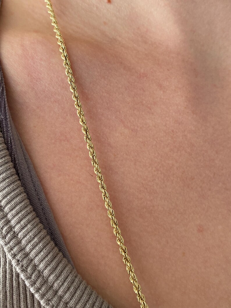 Puede incluir: Un collar de cadena de cuerda de color dorado. La cadena est&aacute; colocada sobre el pecho y la clav&iacute;cula de una persona. El collar est&aacute; hecho de peque&ntilde;os eslabones enrollados, creando una textura similar a la de una cuerda. El fondo es un tono de piel neutro y una camisa gris.