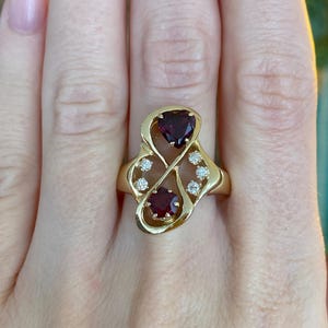 May include: A gold ring with two heart-shaped, deep red gemstones and several small, clear gemstones. The ring has a unique, flowing design.