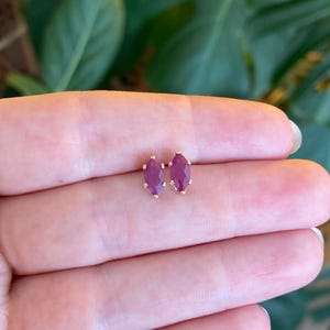 May include: A pair of small stud earrings featuring marquise-cut, deep magenta-colored gemstones set in gold-toned metal prongs. The earrings are positioned side-by-side, showcasing the elongated, pointed shape of the stones and the metallic setting.