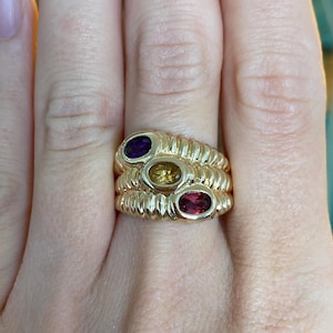 May include: A gold ring featuring three oval gemstones in purple, yellow, and red. The ring has a ribbed design around the gemstones.