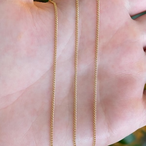 May include: A thin, gold-colored metal chain necklace featuring a delicate, interlocking wheat or spiga link design. The chain is composed of small, uniform, polished links that create a continuous, textured, and flexible strand of jewelry.
