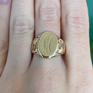 May include: A gold signet ring with an oval face, featuring engraved initials. The ring has decorative detailing on the shoulders. The ring is a warm gold color and is shown on a finger.