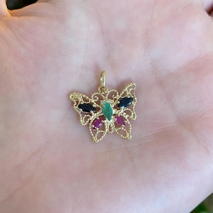 May include: A gold butterfly pendant featuring a filigree wing design. The center of the butterfly is set with a marquise-cut green gemstone, flanked by two dark blue marquise-cut stones on the upper wings and two pink marquise-cut stones below.