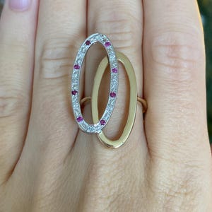 May include: A ring featuring two interlocking oval shapes. One oval is crafted from silver-toned metal and is set with small, clear stones and four vibrant pink stones. The second oval is made of smooth, polished yellow gold-toned metal.
