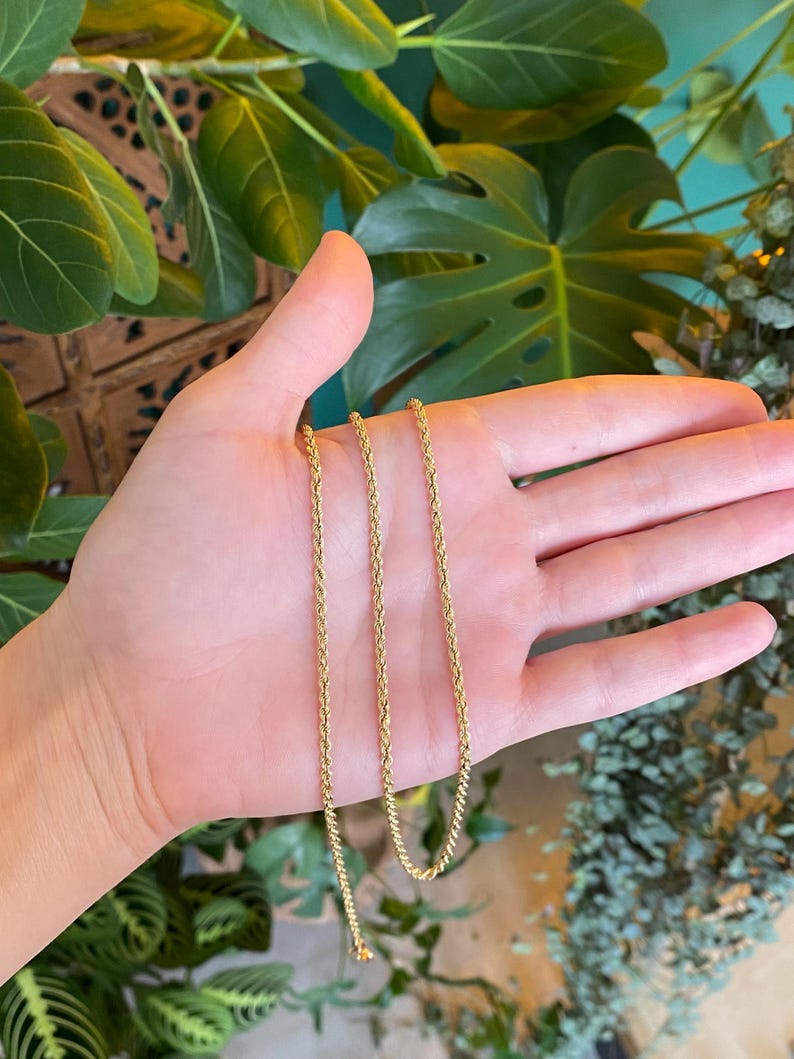 Puede incluir: Un delicado collar de cadena de cuerda de color dorado. La cadena se sostiene en una mano, mostrando sus finos detalles y el tono c&aacute;lido del metal. El fondo presenta un exuberante follaje verde, proporcionando un contraste natural con la joya.