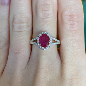 May include: A gold ring with an oval, deep red gemstone encircled by a halo of small, clear stones. The ring's band splits either side of the central stone, forming an open design. The ring is on a person's finger.