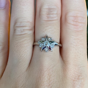 May include: A silver-toned ring featuring a five-pointed star emblem. The star is decorated with colored enamel sections in green, red, blue, and white, accented by small clear stones set between the points of the star on a thin metal band.