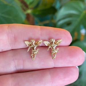 May include: A pair of gold-colored angel stud earrings. Each earring features a detailed cherub design with outstretched wings and delicate features. The earrings are held up against a blurred green background.