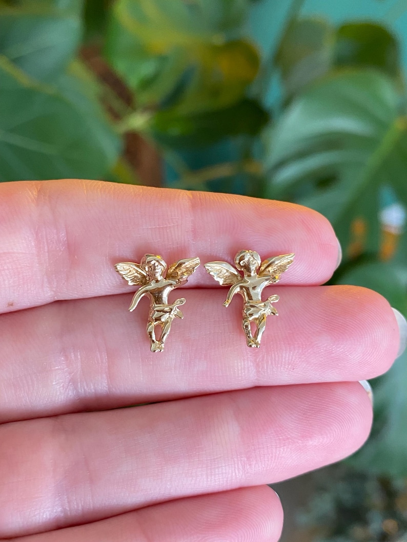 May include: A pair of gold-colored angel stud earrings. Each earring features a detailed cherub design with outstretched wings and delicate features. The earrings are held up against a blurred green background.
