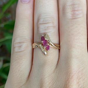 May include: A gold ring featuring a bypass design with three marquise-cut red gemstones set vertically in the center. The polished gold band curves around the stones, creating a sleek and continuous look that highlights the vibrant red color.