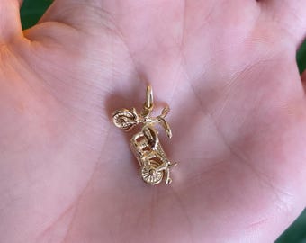 May include: A small, gold-colored scooter charm. The charm features intricate details of a vintage scooter, including handlebars, a seat, and wheels. The charm is designed to be a pendant or a charm for a bracelet.