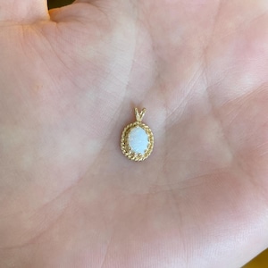 May include: A gold pendant with an oval, light-colored stone. The stone is surrounded by a twisted rope design. The pendant is held in a hand, showing the scale of the jewelry.