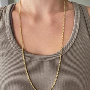 Puede incluir: Un collar largo de cadena de cuerda dorada. La cadena est&aacute; drapeada alrededor del cuello y cuelga sobre el pecho. El collar se usa sobre una camiseta sin mangas de color neutro.